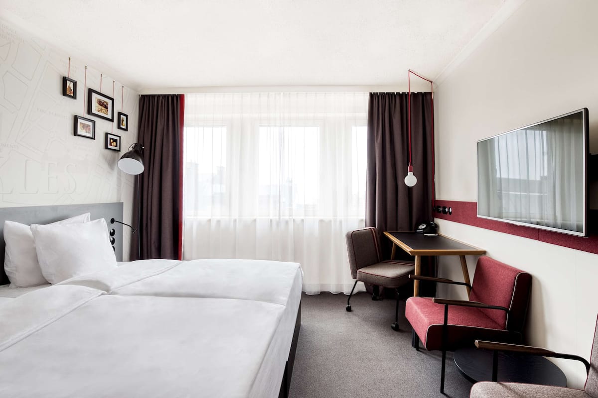 Une chambre d’hôtel avec un lit, un bureau, une chaise, une télévision et une fenêtre avec des rideaux.