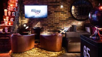 Eine gemütliche Lounge mit Ledersofas, einem Fernseher, der 'NEED SPEED' zeigt, und einer Ziegelwand.