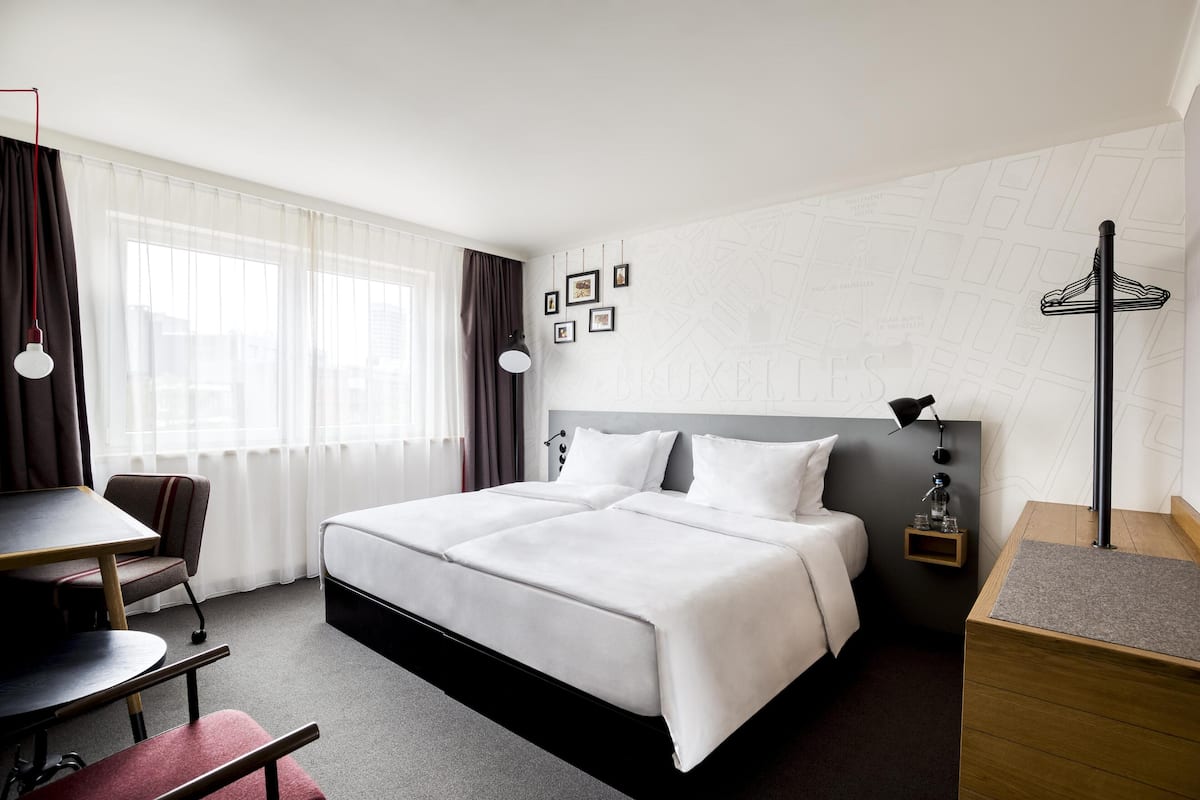 Une chambre d’hôtel avec un grand lit, un bureau et une chaise. On y trouve des rideaux, une lampe et une petite étagère avec une bouteille.