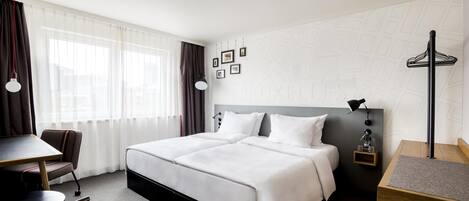 A hotel room with a large bed, a desk, and a chair. There are curtains, a lamp, and a small shelf with a bottle.