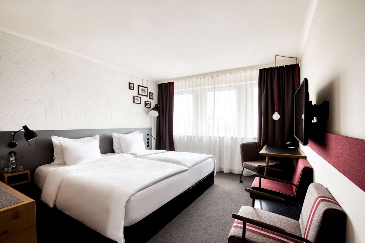 Ein Hotelzimmer mit einem großen Bett, einem Schreibtisch, einem Sessel, einem Fernseher und einem Fenster mit Vorhängen.
