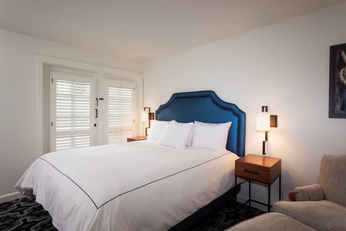 A hotel room with a large bed, two bedside tables, a chair, and a door with white blinds.