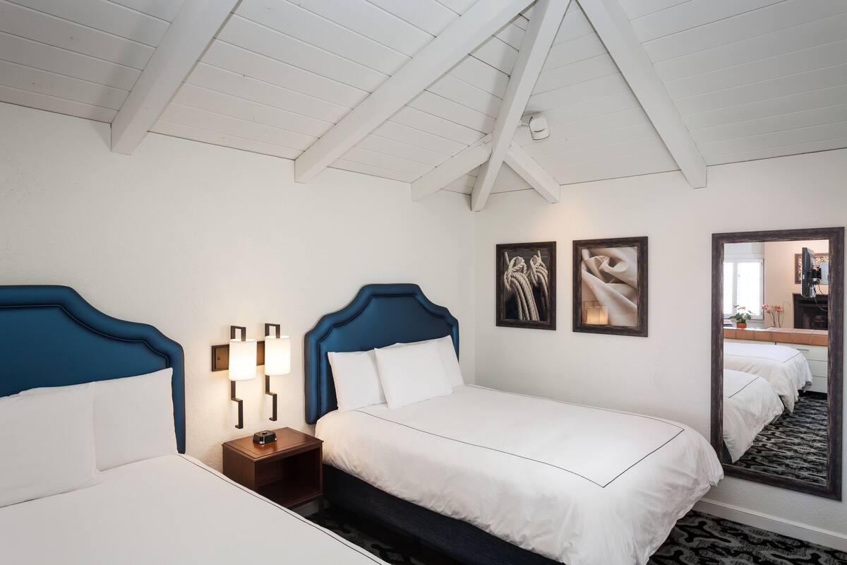 A hotel room with two beds, white bedding, a wooden headboard, a nightstand with a lamp, and a large mirror.