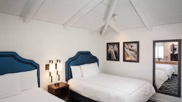 Habitación de hotel con dos camas, ropa de cama blanca, cabecero de madera, mesita de noche con lámpara y un espejo grande.