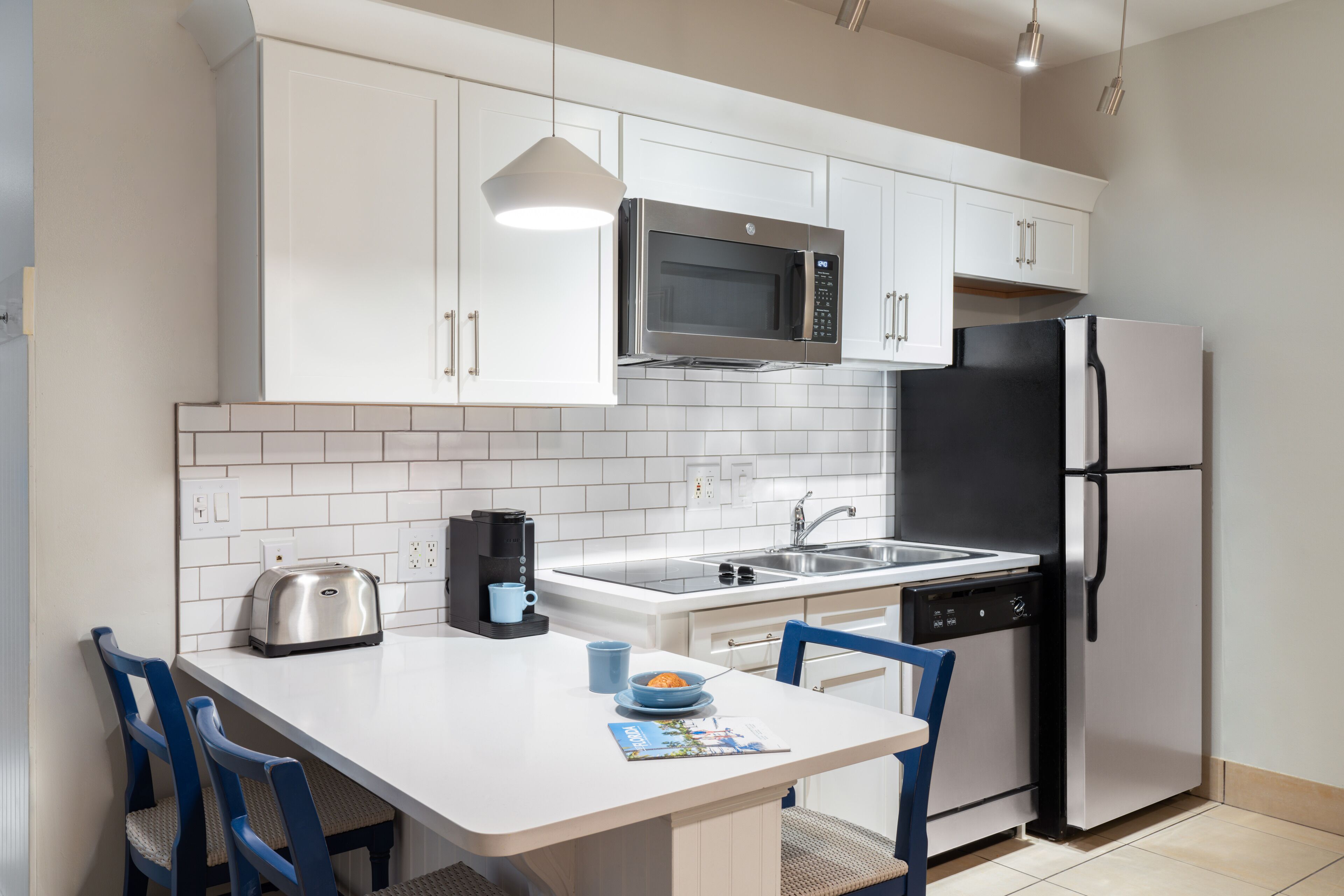 A modern kitchen with white cabinets, a stainless steel microwave, a black refrigerator, a sink, and a dining area with blue chairs.