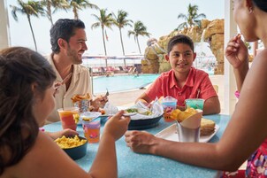 A group of people dining at a poolside table with a view of palm trees and a rocky backdrop.