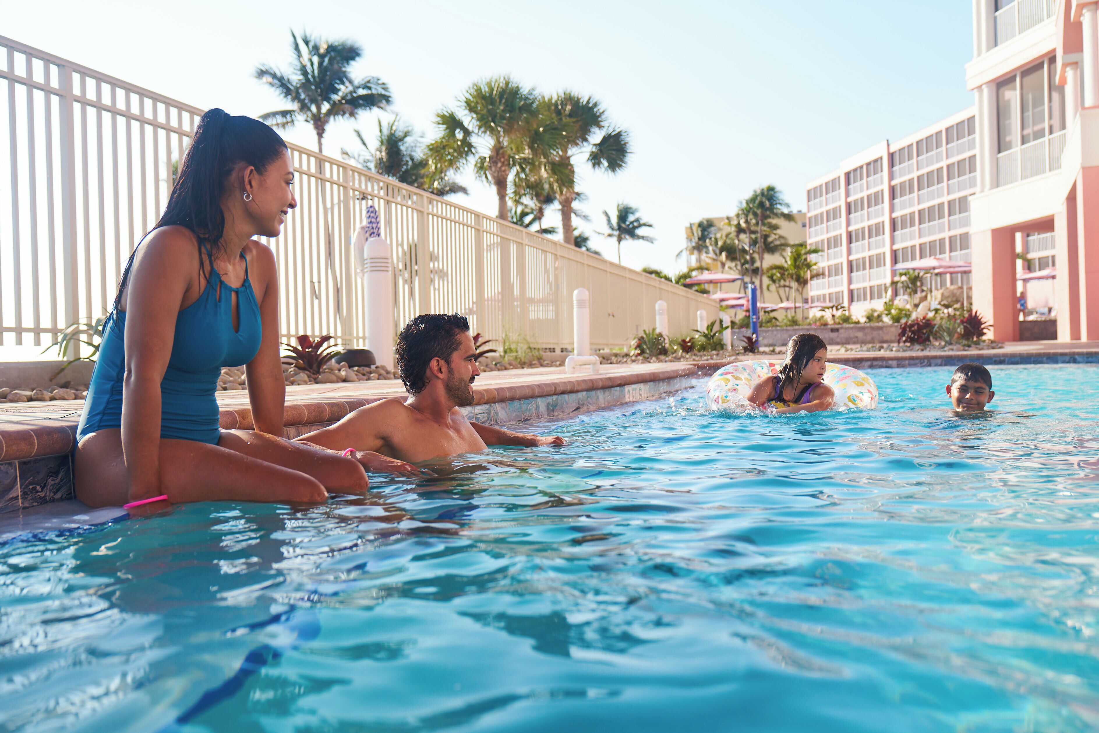 Zona de piscina con gente relajándose, una valla junto a la piscina y palmeras al fondo.