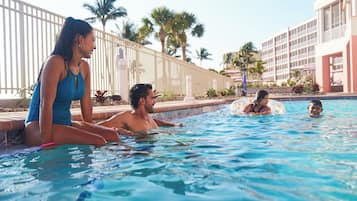 Zona de piscina con gente relajándose, una valla junto a la piscina y palmeras al fondo.