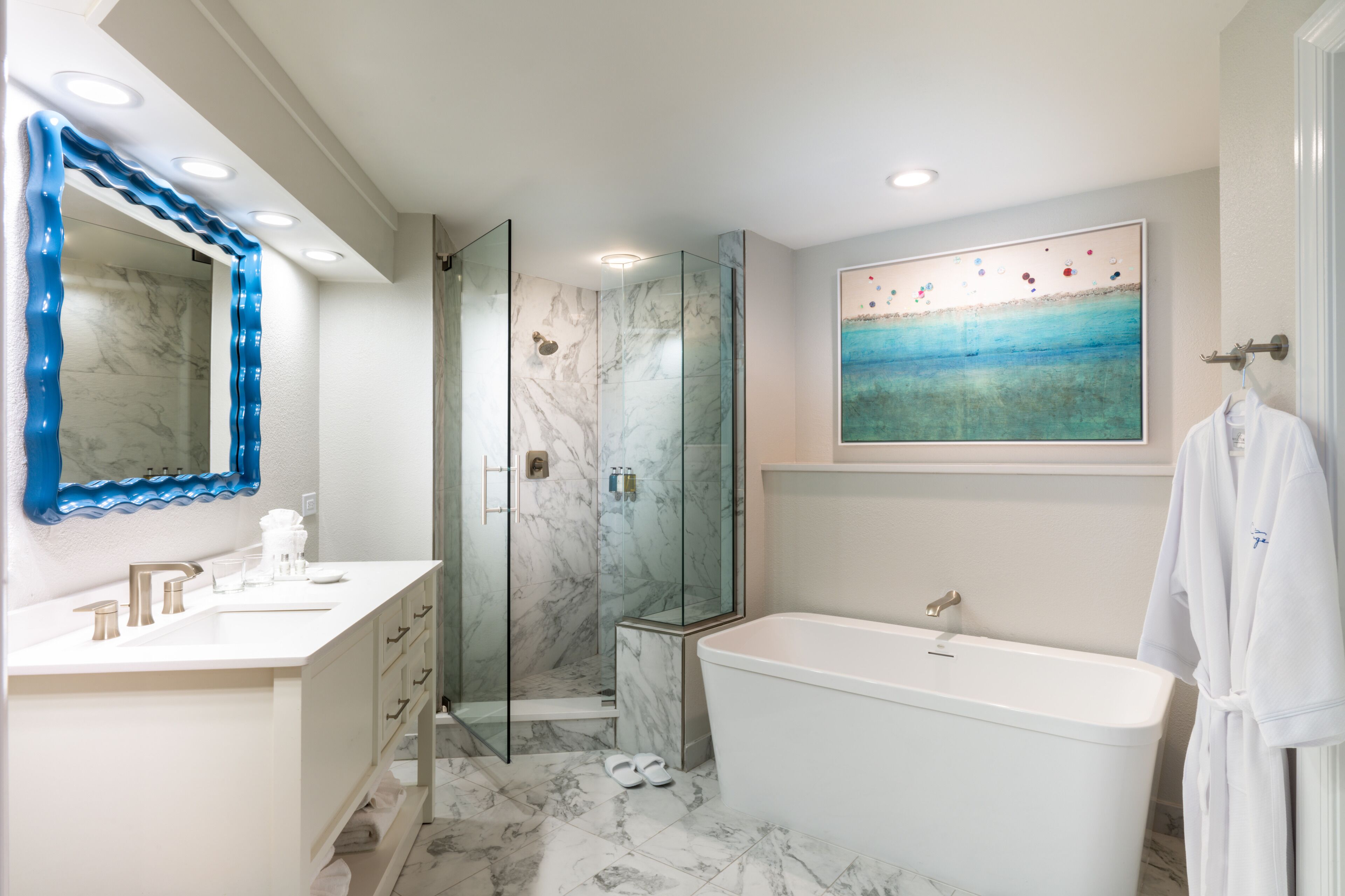 A modern bathroom with a marble vanity, a glass-enclosed shower, a large bathtub, and a beach-themed artwork on the wall.