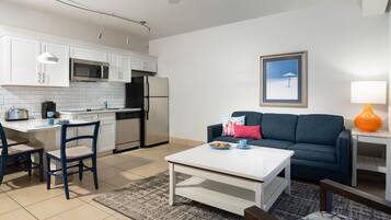A modern kitchen with white cabinets, a microwave, and a refrigerator. There is a dining area with a white table and blue chairs. A blue sofa and a framed picture are in the living area.