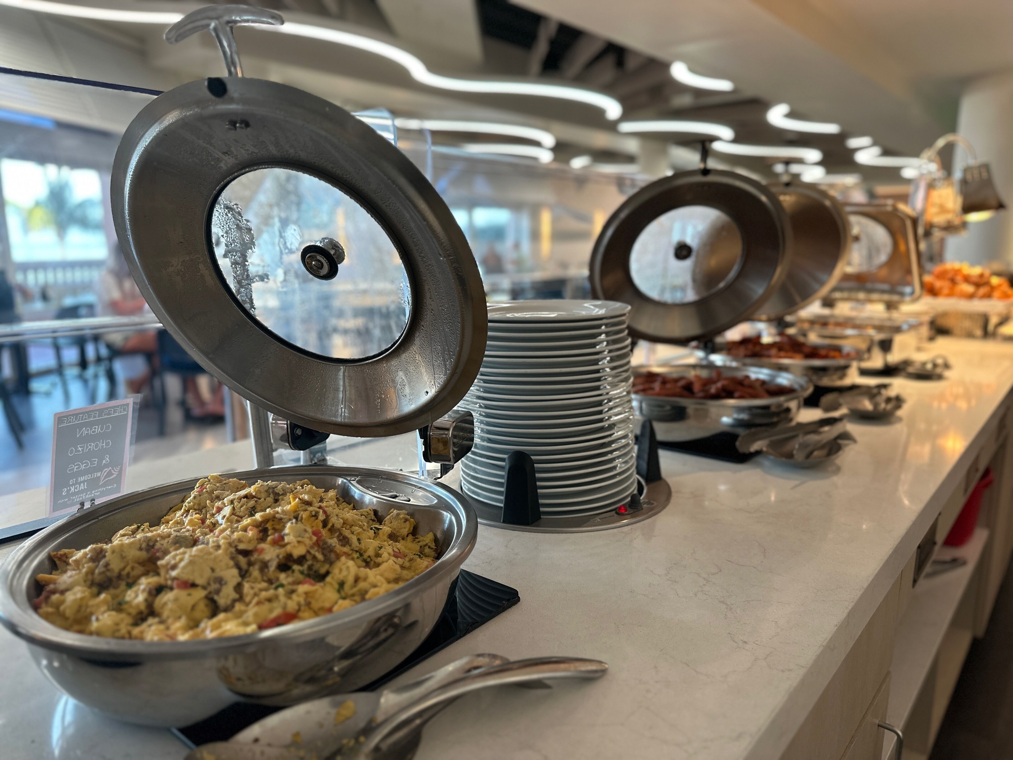 A buffet with various dishes, including scrambled eggs and meats, arranged on a counter with stacked plates and serving utensils.
