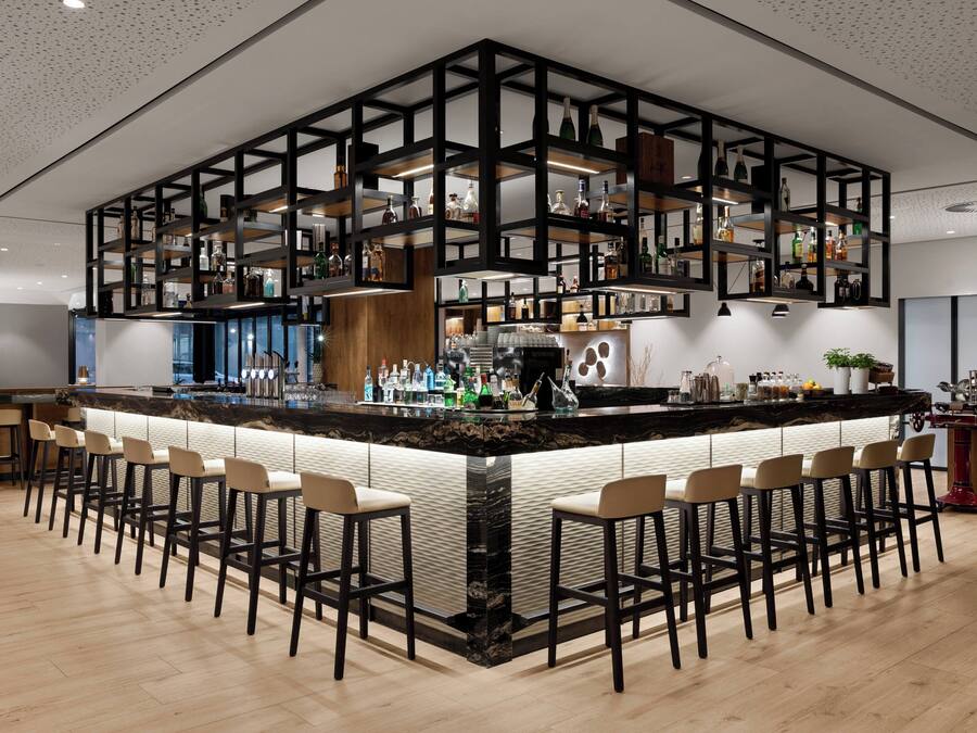 Una zona de bar moderna con una barra central, diversas opciones de asientos y una variedad de botellas y vasos en estanterías.