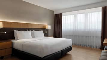Une chambre d’hôtel moderne avec un grand lit, une tête de lit en bois et une vue sur la ville à travers de fines rideaux.