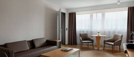 A modern hotel room with a brown leather sofa, a wooden coffee table, a round wooden side table, two armchairs, a small round table with a plant, and a large window with curtains.