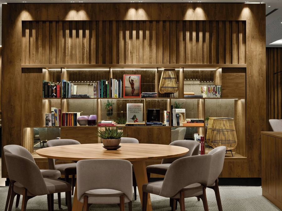 Una biblioteca moderna con un panel de madera, estanterías, una mesa redonda de madera y asientos cómodos.