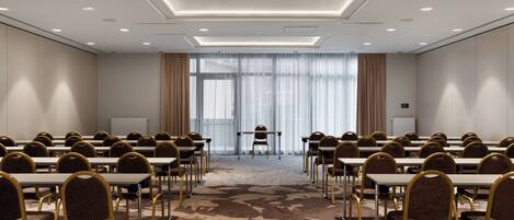 Un salón de conferencias con filas de mesas y sillas, ventanas amplias con cortinas y suelo alfombrado.