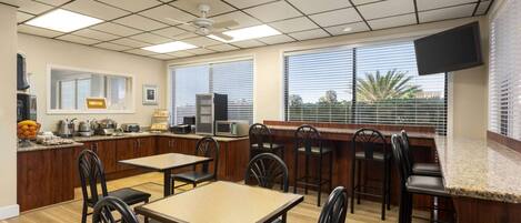 A restaurant with a bar area, multiple tables and chairs, a kitchen counter with appliances, and large windows with blinds.