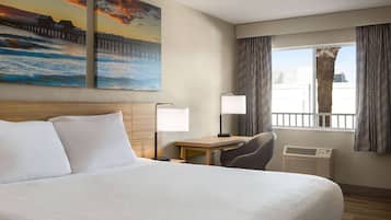 Une chambre d’hôtel avec un grand lit, un bureau, une chaise, une fenêtre avec des rideaux et deux tableaux accrochés au mur.