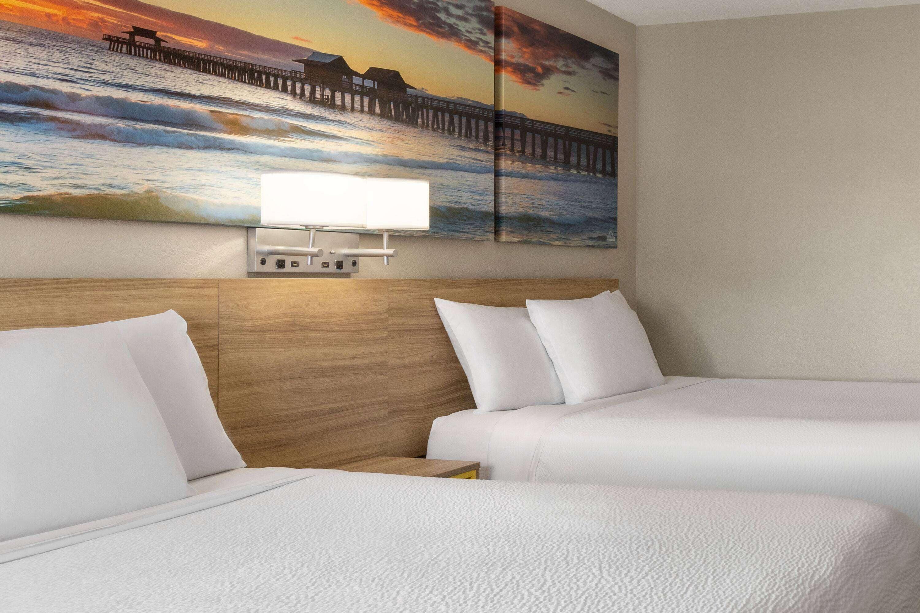 Une chambre d’hôtel avec deux lits, une tête de lit en bois, un tableau représentant une jetée et un lampadaire fixé au mur.