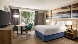 A hotel room with a bed, a desk, a chair, a table, a TV, and a painting of a sunset over a pier. - Days Inn by Wyndham Cocoa Beach Port Canaveral (Cocoa Beach)
