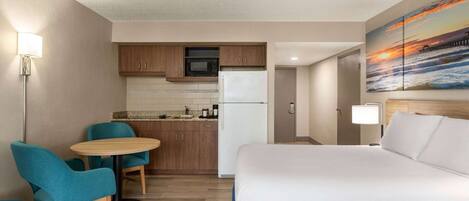 Habitación de hotel con cama, kitchenette pequeña, zona de comedor con sillas y un cuadro de una puesta de sol sobre el océano.