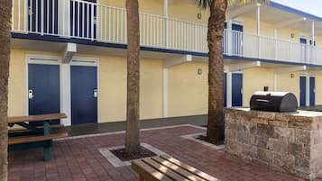 Un edificio de dos plantas con balcones y barandillas blancas, una parrilla en una plataforma de piedra y una mesa de picnic en la planta baja.