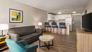 A modern living room with a grey sofa, teal armchair, and a small round coffee table. A kitchen area with white cabinets and bar stools is visible in the background. - Days Inn by Wyndham Cocoa Beach Port Canaveral (Cocoa Beach)