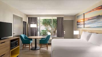 A hotel room with a bed, a TV, a desk with two chairs, a window with curtains, and a painting of a sunset.
