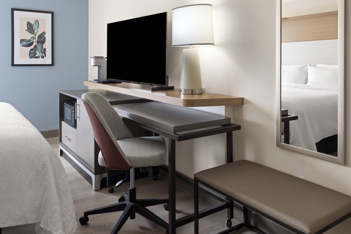 A hotel room with a bed, a desk with a chair, a television, a lamp, and a large mirror.
