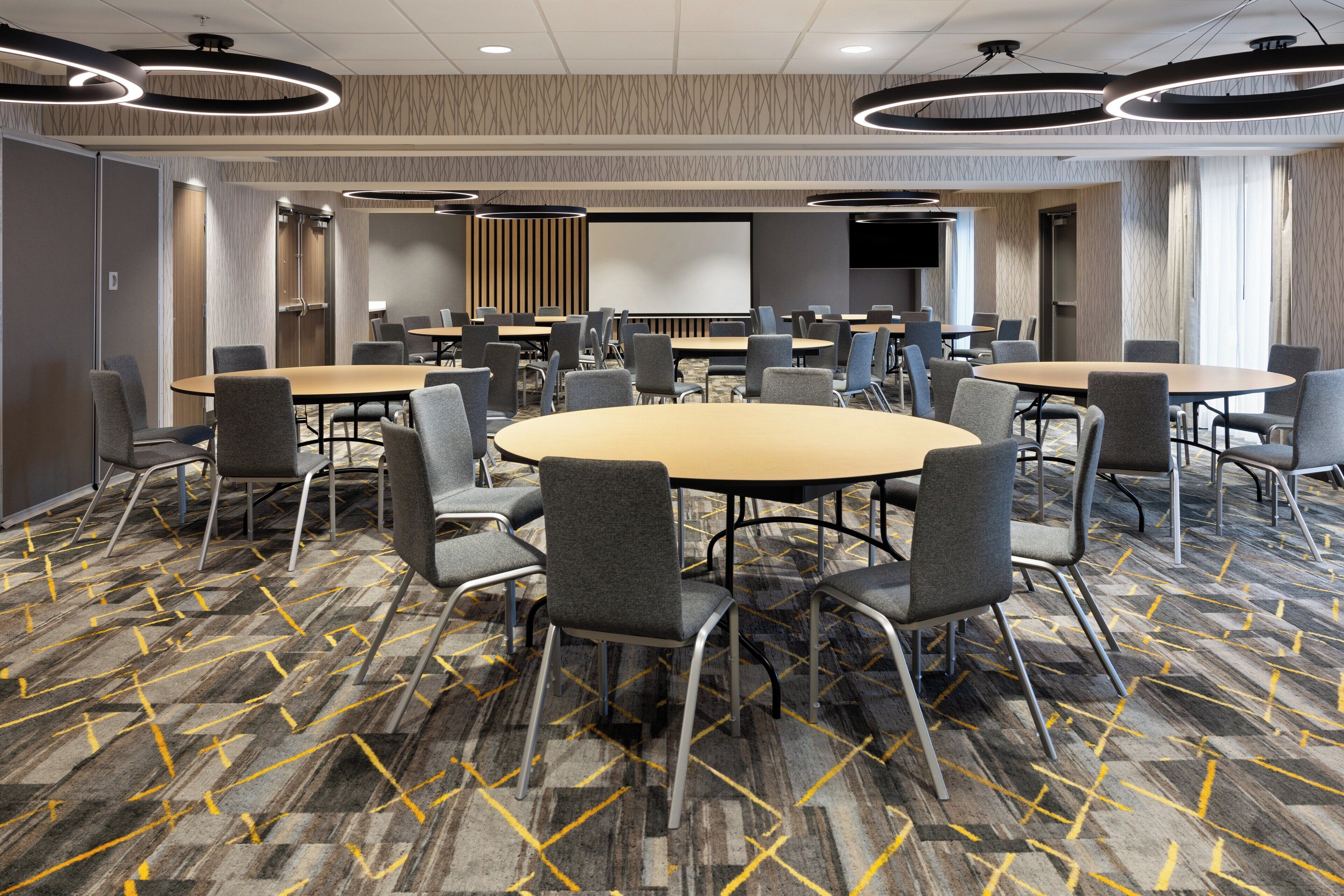 A conference room with multiple tables and chairs, a large screen, and a patterned carpet.