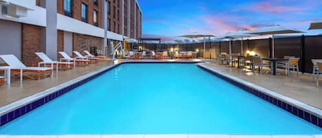 A swimming pool with lounge chairs and tables, surrounded by a brick building with large windows.