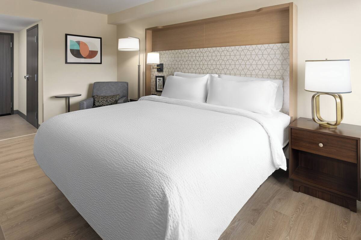 A hotel room with a large bed, a bedside table with a lamp, a chair, and a small table.