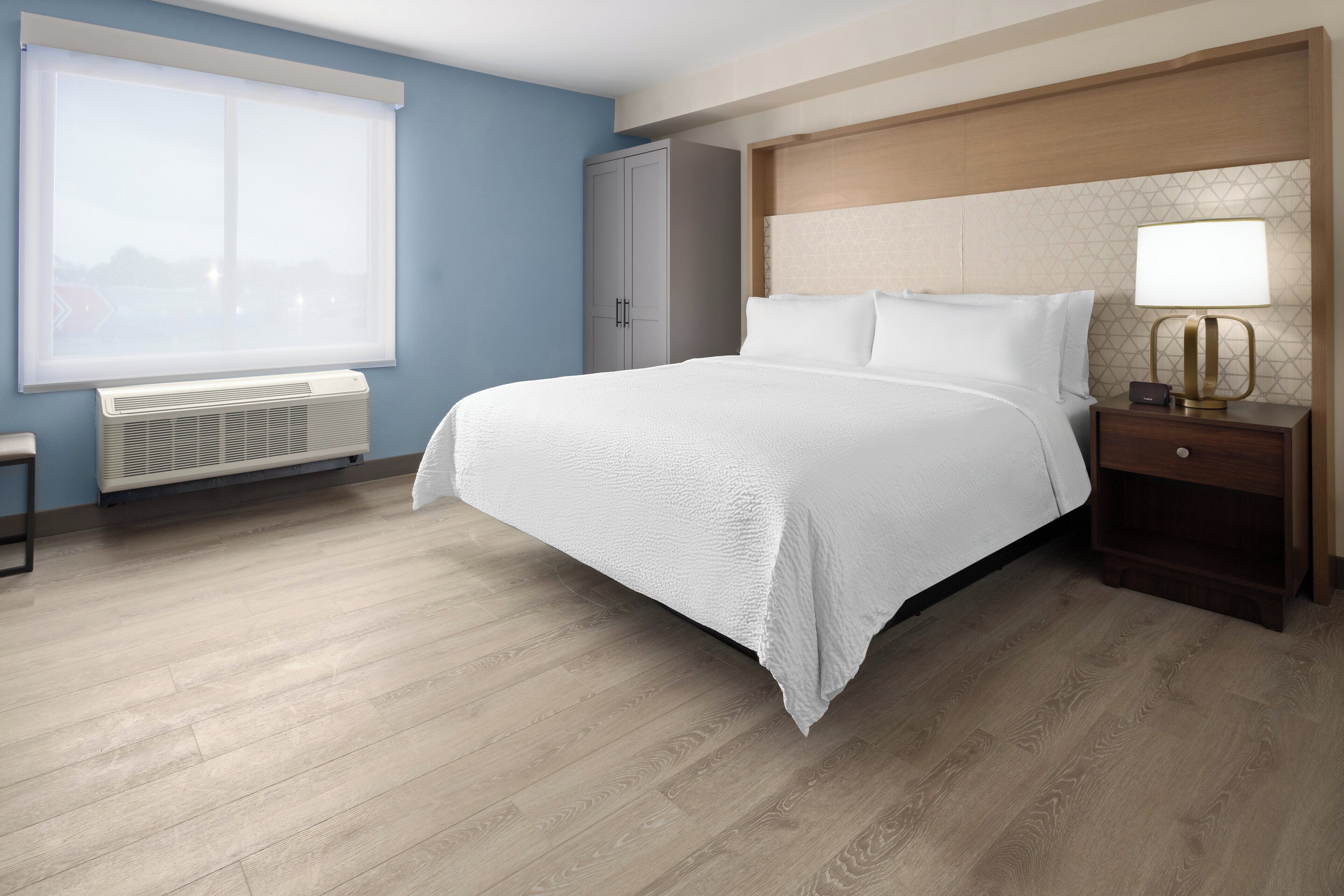 Habitación de hotel con una cama grande, una mesita de noche con lámpara, un armario y una ventana con persianas.