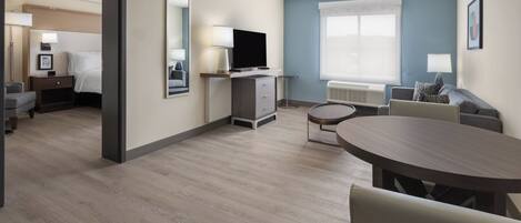 Une chambre d’hôtel comprenant un lit, un bureau avec une télévision, un coin repas avec une table et des chaises, et une fenêtre avec des stores.