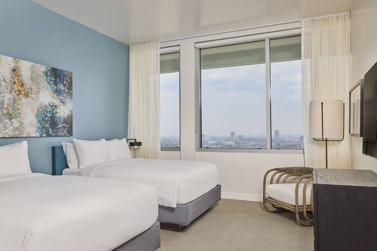 Habitación de hotel con dos camas, vista a la ciudad, un cuadro abstracto grande y una silla de mimbre.