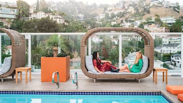 A rooftop pool area with two wicker lounge chairs, a potted plant, and a view of a hillside residential area.