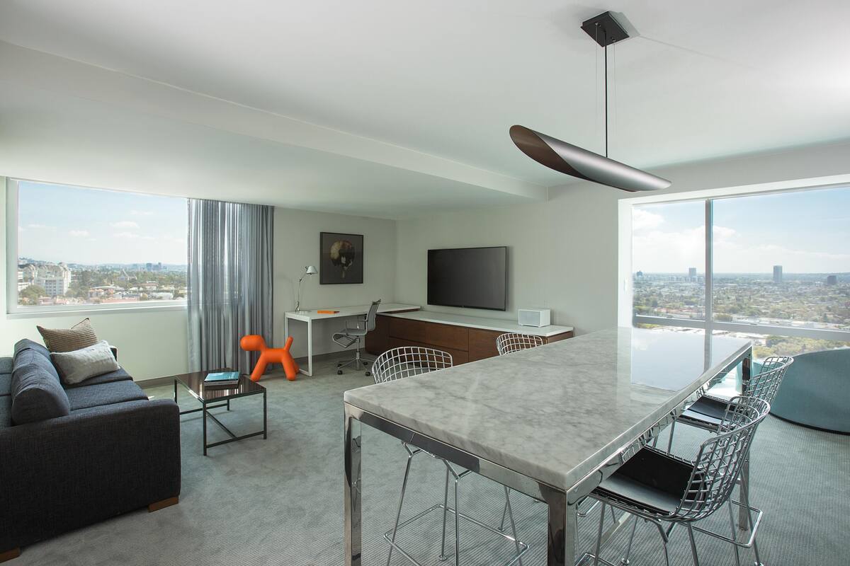 Una habitación de hotel moderna con un ventanal grande, una zona de comedor, un sofá, un escritorio y un televisor.