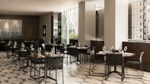 A modern restaurant interior with multiple dining tables and chairs, a patterned floor, and a large window providing natural light.