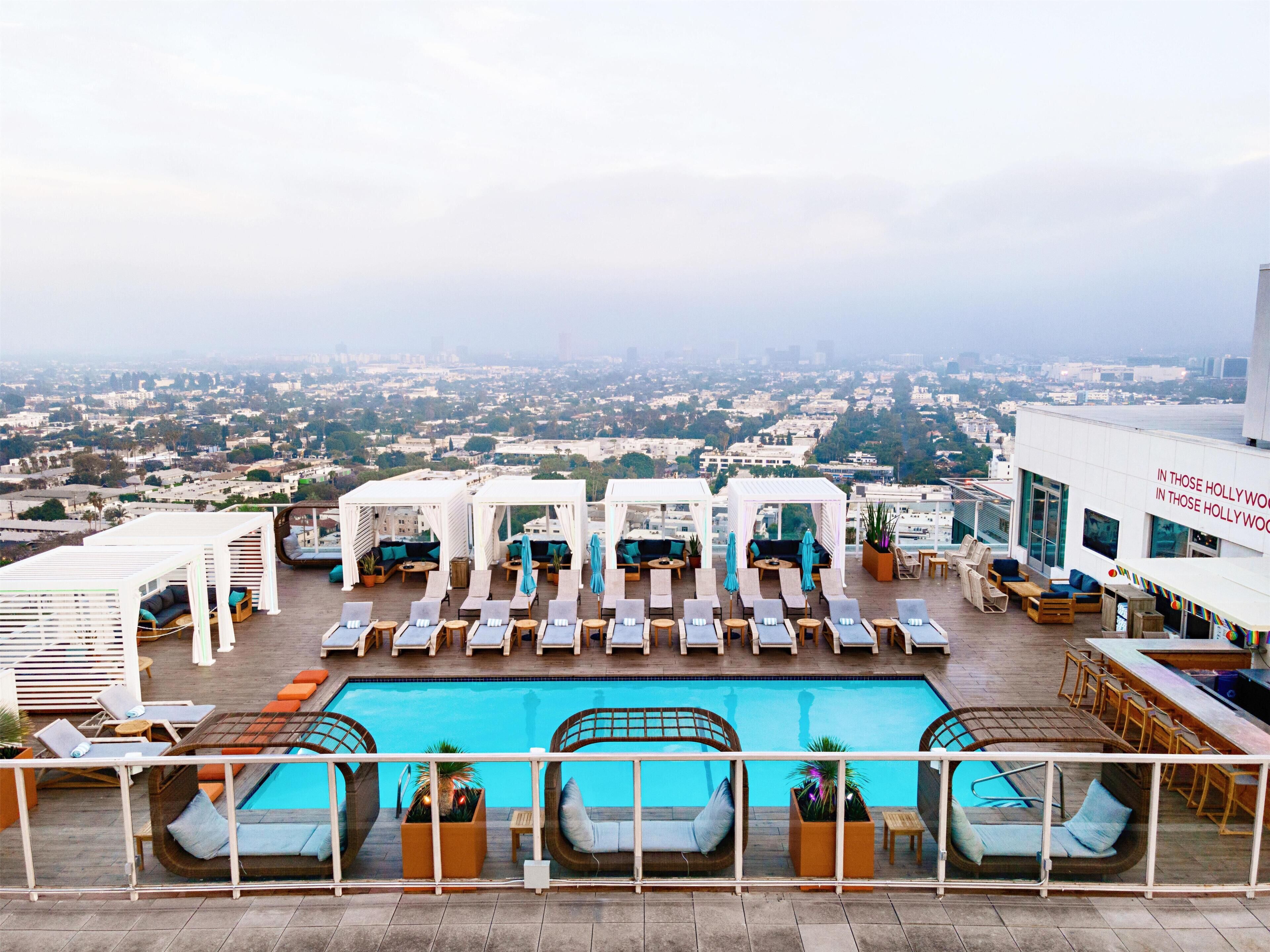 A rooftop pool area with lounge chairs and cabanas, overlooking a cityscape.