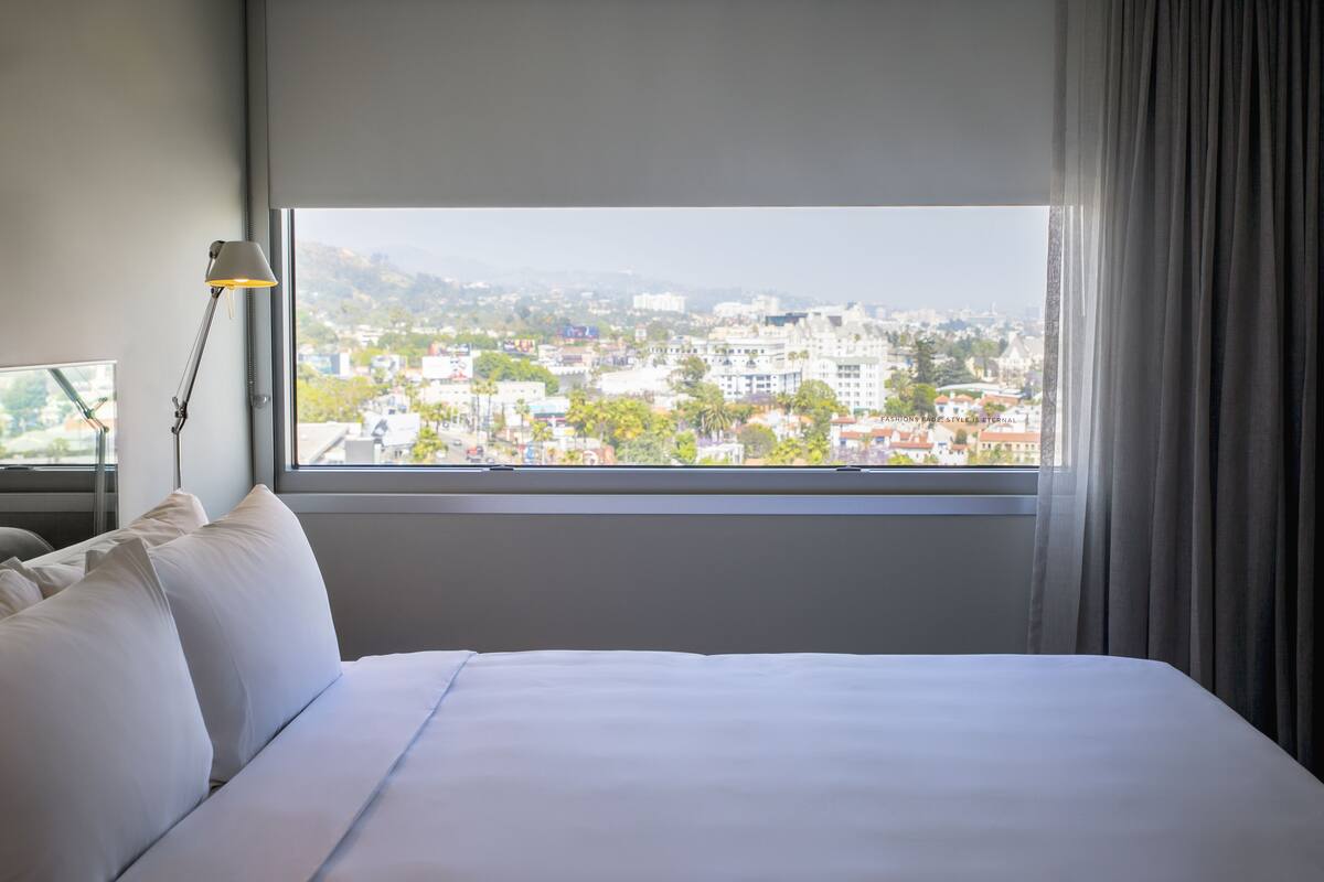 Habitación de hotel con un ventanal grande, una cama con sábanas blancas, una lámpara de escritorio y vistas a un paisaje urbano.