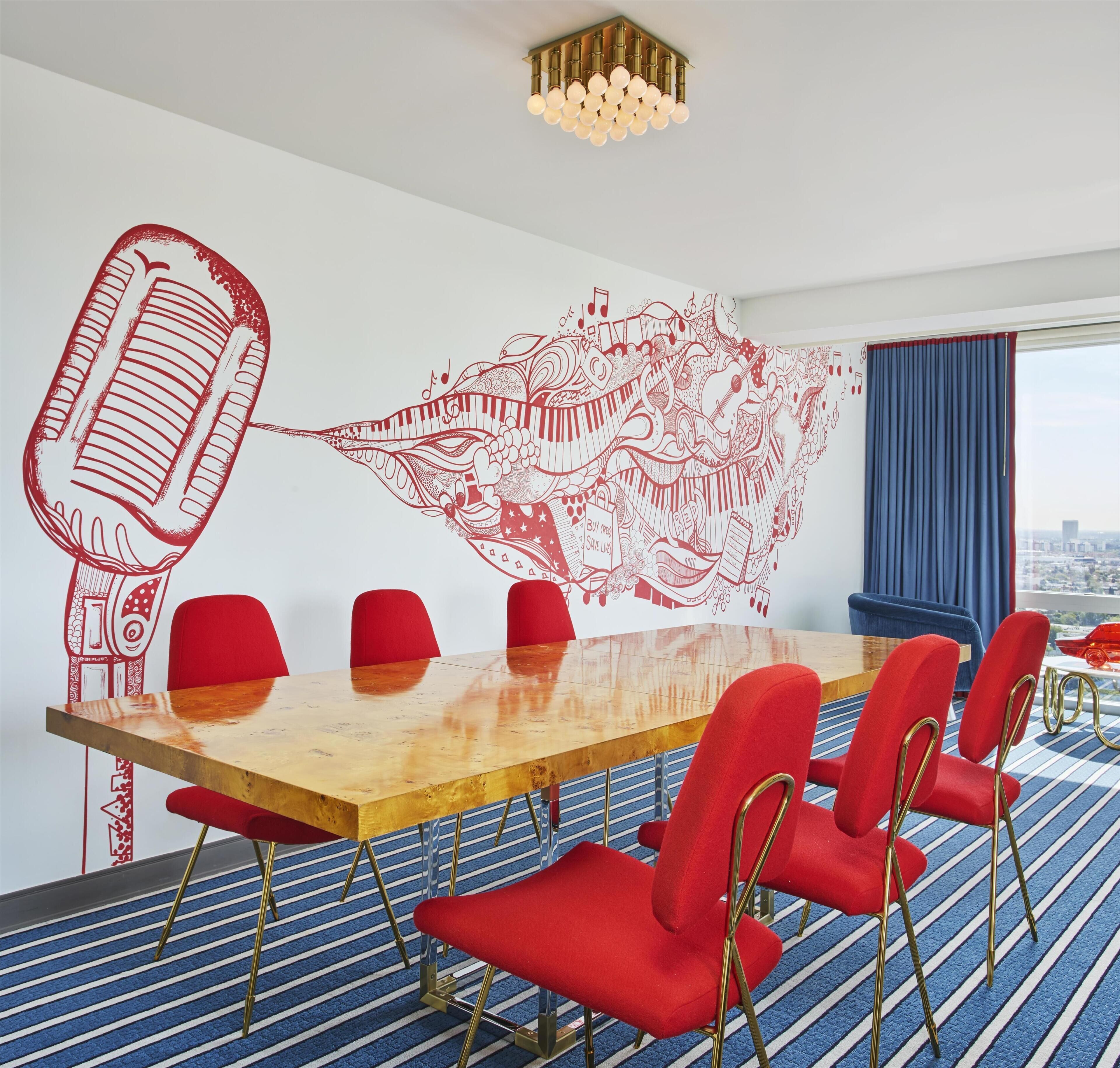 A conference room with a large wooden table, red chairs, a microphone illustration on the wall, and a city view through the window.