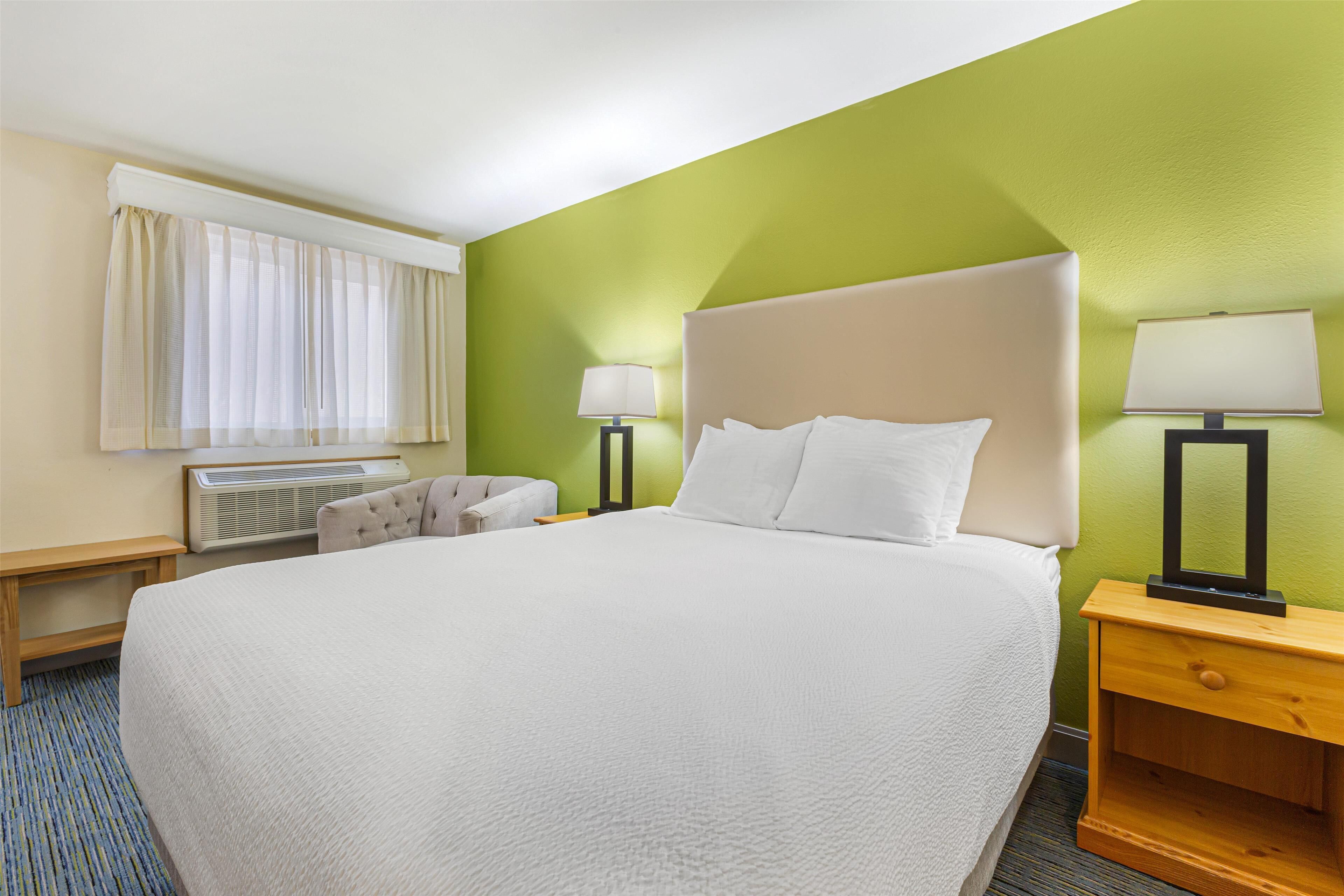Een hotelkamer met een groot bed, een nachtkastje met een lamp, een stoel en een raam met gordijnen.