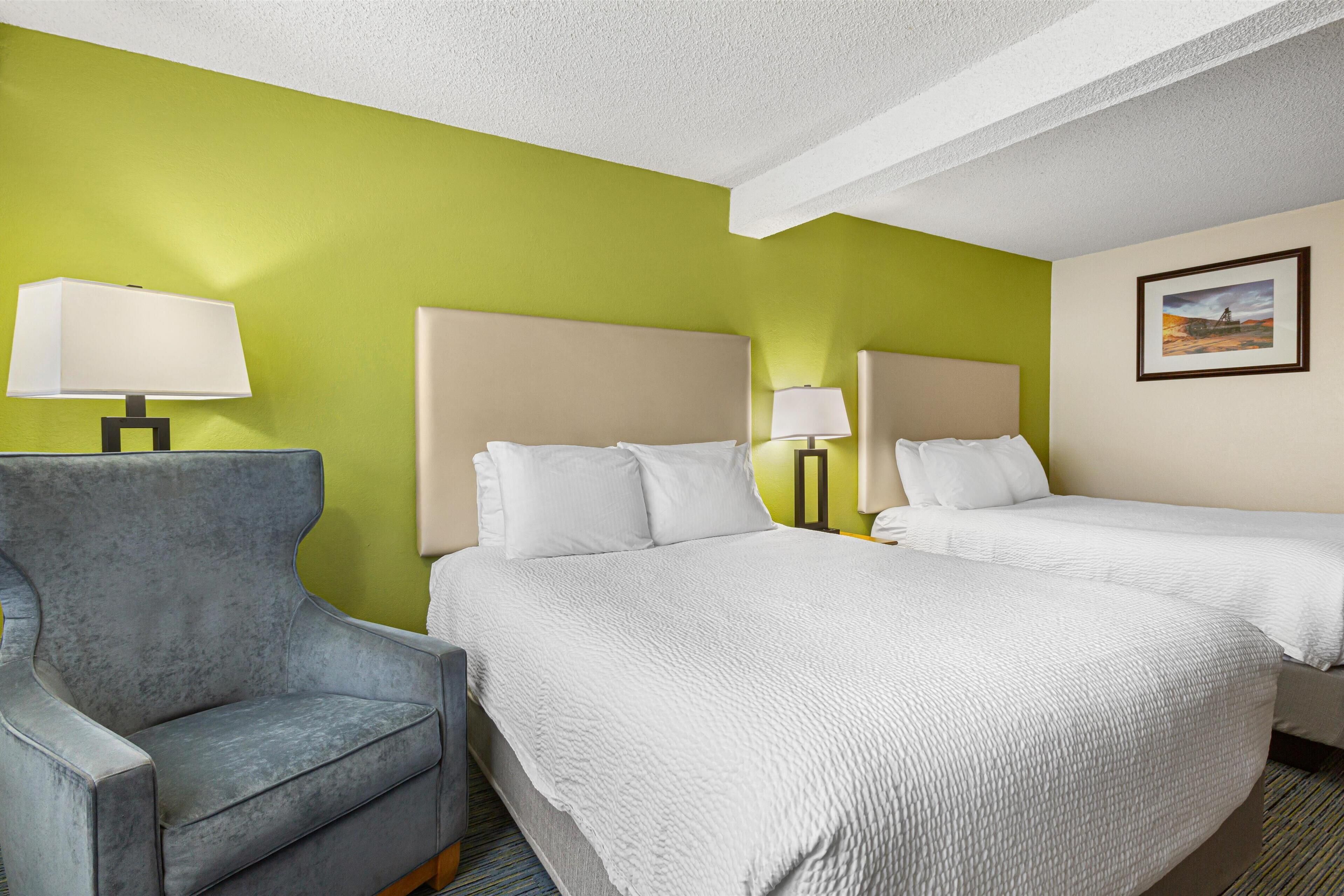 A hotel room with two beds, a grey armchair, and a green wall.