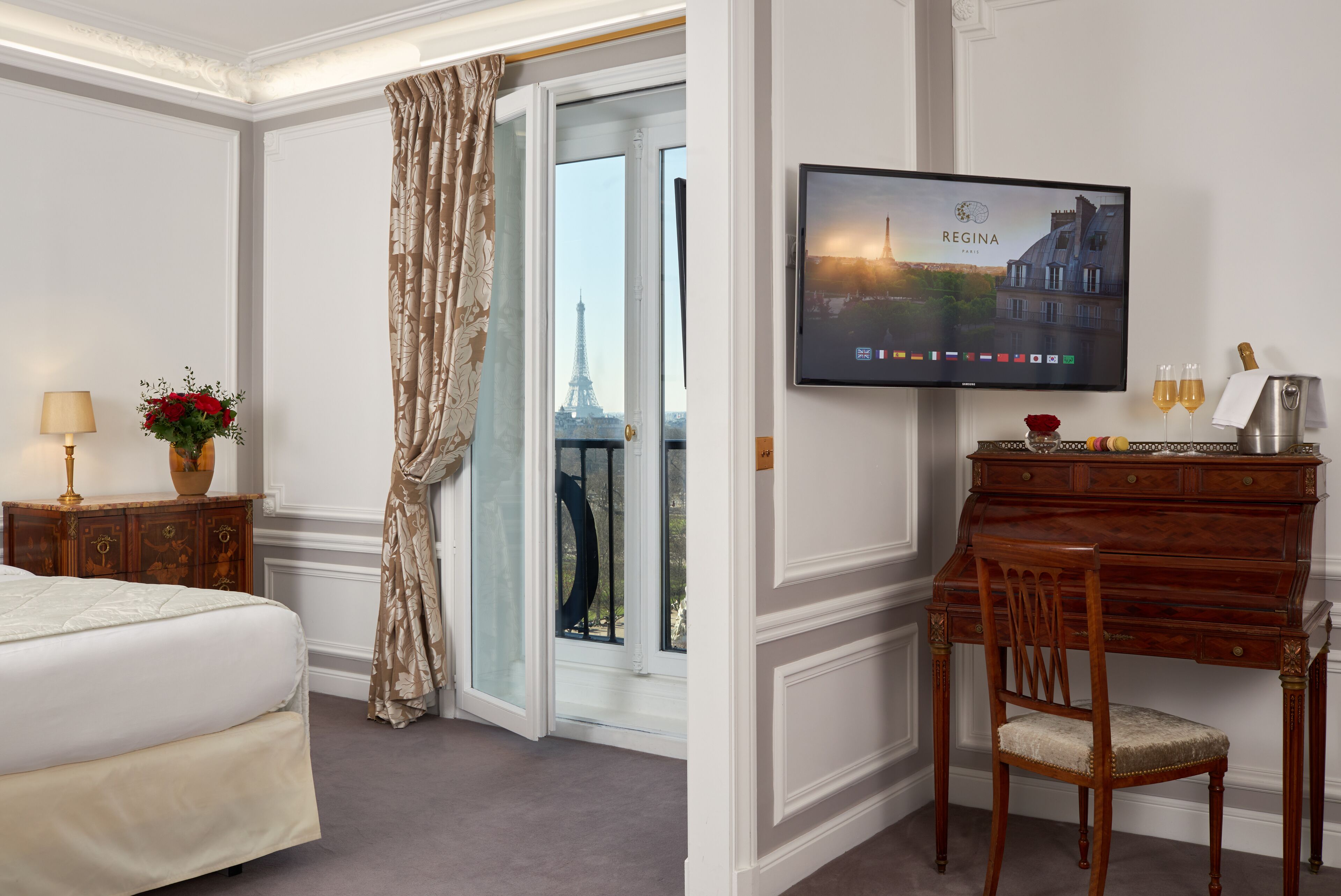 Ein Hotelzimmer mit Bett, Schreibtisch mit Stuhl, Fernseher der eine Stadtansicht zeigt und Blick auf den Eiffelturm durch eine Glastür.