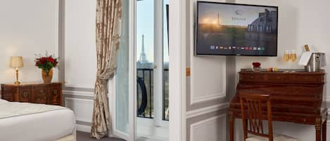 Una habitación de hotel con una cama, un escritorio con una silla, un televisor mostrando un paisaje urbano y vistas de la Torre Eiffel a través de una puerta de cristal.