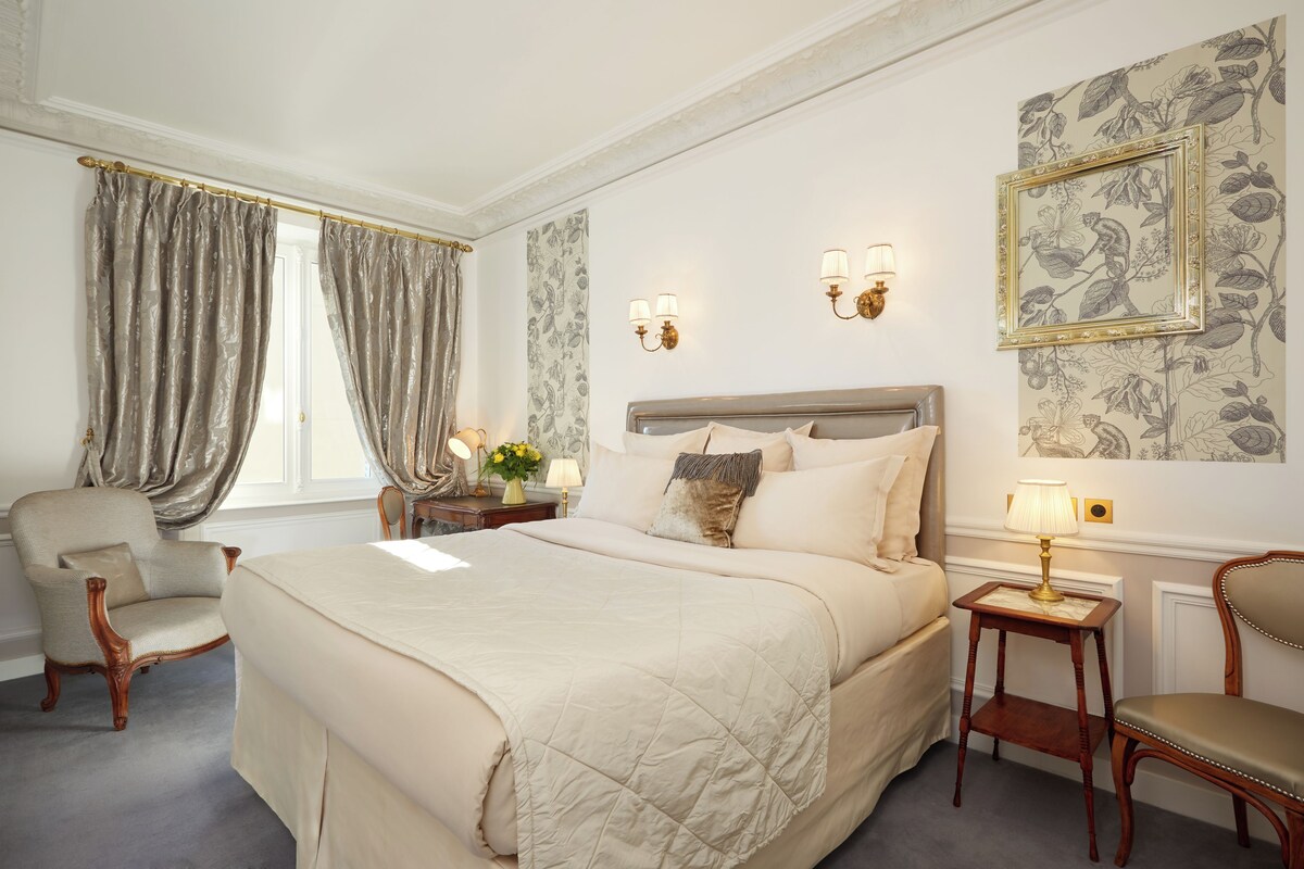 Een slaapkamer met een groot bed, twee stoelen, een nachtkastje, een lamp en een bloemen behang.
