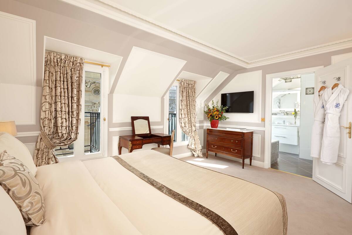 A spacious bedroom with a large bed, a dresser, a TV, and a bathroom visible through an open door.