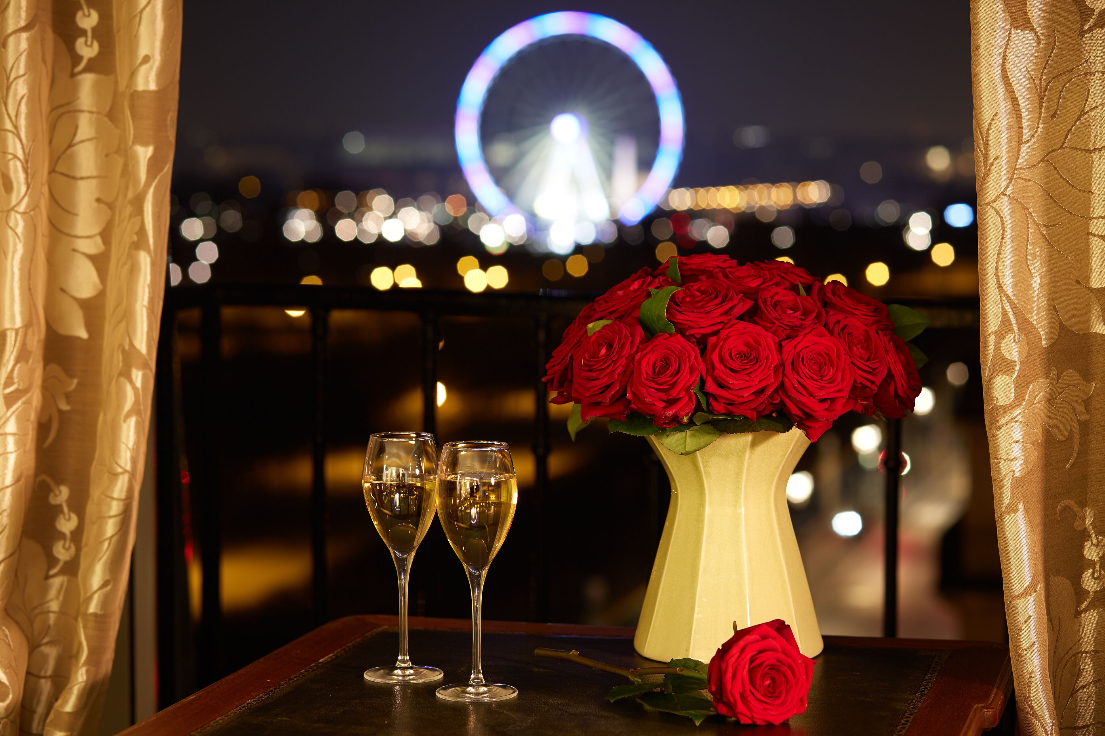 A table with a vase of red roses and two glasses of champagne, with a Ferris wheel in the background.