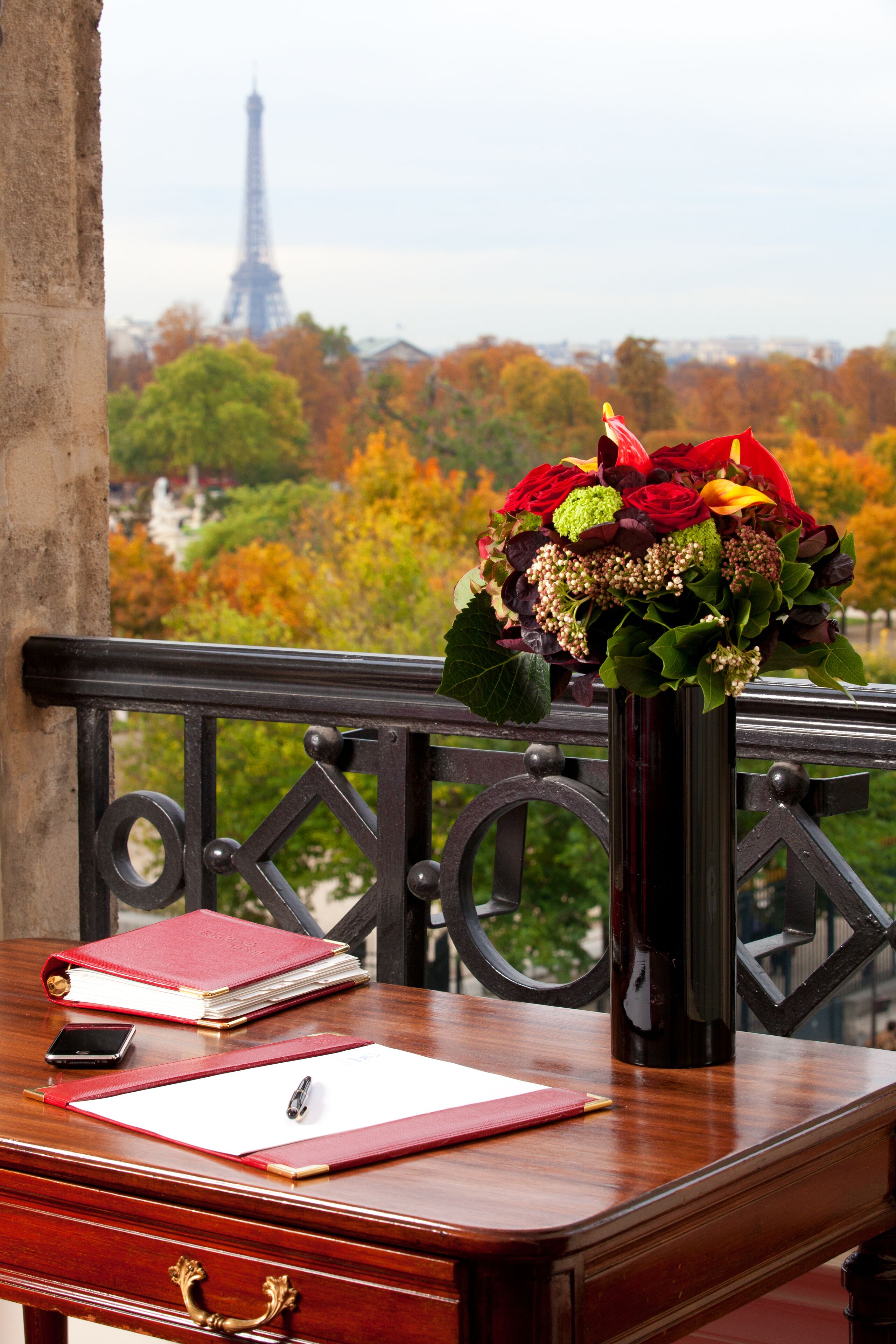 Una scrivania in legno con una cartella rossa, una penna e uno smartphone. Sul tavolo c'è un vaso con dei fiori. Sullo sfondo, si vede una vista di alberi e la Torre Eiffel.