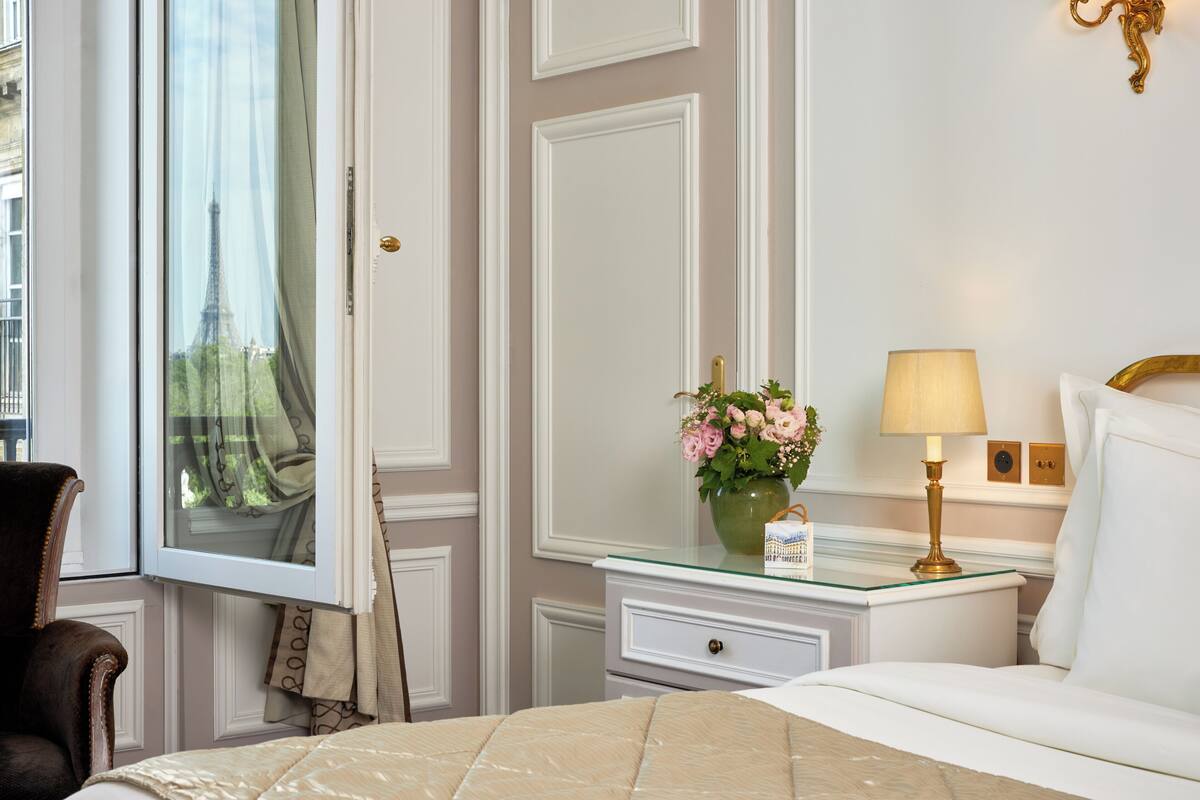 A hotel room with a bed, a nightstand with a lamp, a vase with flowers, a window with curtains, and a view of the Eiffel Tower.